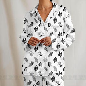 K&ouml;nnte beinhalten: Wei&szlig;es Pyjama-Set mit einem sich wiederholenden Muster von Cartoon-B&ouml;sewicht-Gesichtern in Schwarz und Grau. Das lang&auml;rmlige Oberteil hat einen Kragen und eine Knopfleiste. Die Pyjamahose ist ebenfalls sichtbar.