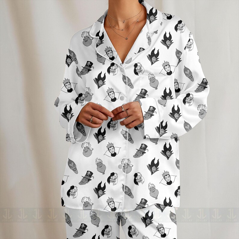 K&ouml;nnte beinhalten: Wei&szlig;es Pyjama-Set mit einem sich wiederholenden Muster von Cartoon-B&ouml;sewicht-Gesichtern in Schwarz und Grau. Das lang&auml;rmlige Oberteil hat einen Kragen und eine Knopfleiste. Die Pyjamahose ist ebenfalls sichtbar.
