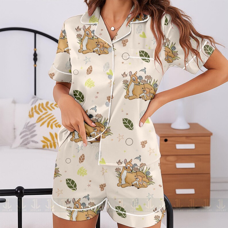 Op de afbeelding: Cr&egrave;mekleurige pyjamaset met een Bambi en Stampertje print. De top met korte mouwen en de shorts zijn versierd met een herhalend patroon van de karakters, bloemen, bladeren en sterren. Witte biezen omlijnen de randen van de set.