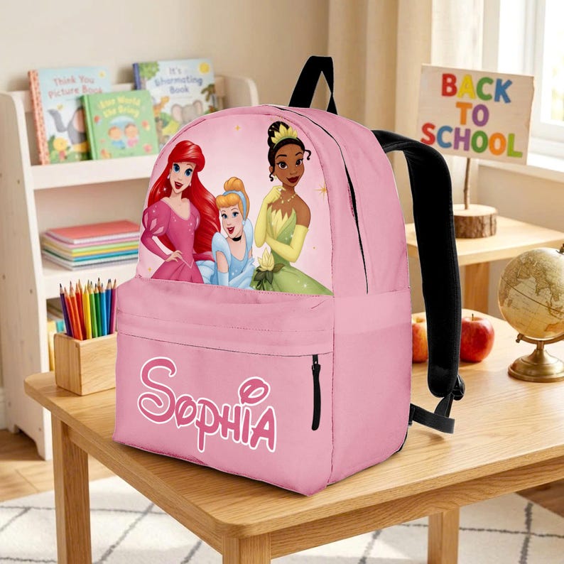 May include: A pink backpack featuring a graphic print of three animated princesses on the front pocket area. The name Sophia is printed in large white letters with a pink outline on the lower front pocket. The bag has black straps and zippers.