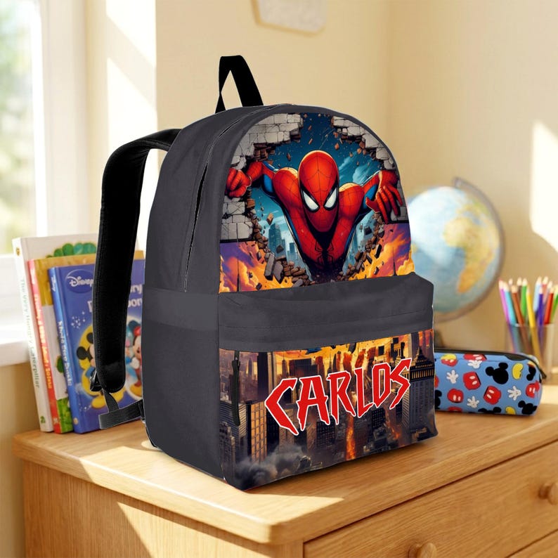 May include: A black backpack featuring a graphic of the superhero Spider-Man breaking through a brick wall on the top section. The bottom pocket displays a city skyline with the name CARLOS printed in a bold, red, jagged comic book font.