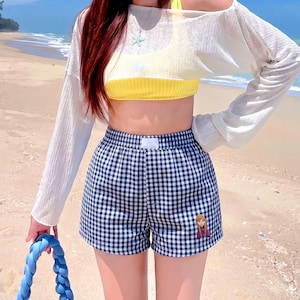 May include: A yellow ribbed crop top paired with a white sheer long-sleeve off-the-shoulder top and black and white gingham shorts. The shorts feature a small embroidered character patch on the lower left leg and a white rectangular waist label.