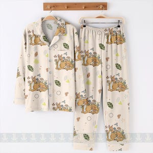 Op de afbeelding: Cr&egrave;mekleurige pyjamaset met een hert- en bloemenprint. De button-down shirt met lange mouwen en de bijpassende broek hebben een herhalend patroon van herten, bloemen, bladeren en vogels. De pyjama hangt aan een houten rek.