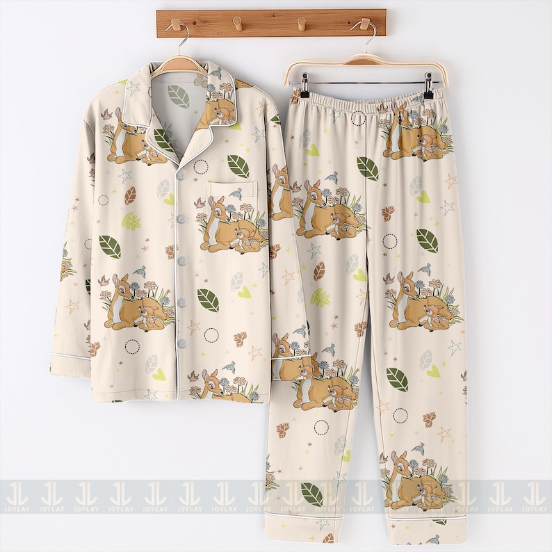 Op de afbeelding: Cr&egrave;mekleurige pyjamaset met een hert- en bloemenprint. De button-down shirt met lange mouwen en de bijpassende broek hebben een herhalend patroon van herten, bloemen, bladeren en vogels. De pyjama hangt aan een houten rek.