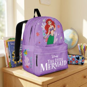 May include: A purple backpack featuring Ariel from Disney's The Little Mermaid. The backpack has a front pocket with a zipper and a large image of Ariel. The words "The Little Mermaid" are printed in white on the front.