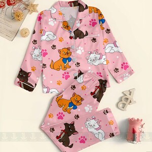 May include: Pink pajama set featuring cartoon cats, paw prints, and white trim. The long-sleeved top has a button-down front and a chest pocket. The matching pants complete the set. The pajama set is made of a soft, comfortable material.