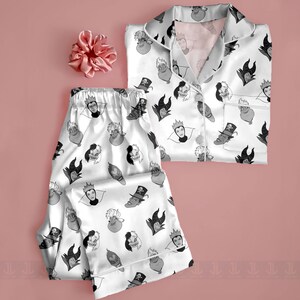 K&ouml;nnte beinhalten: Wei&szlig;es Satin-Pyjama-Set mit einem sich wiederholenden Muster von Zeichentrick-B&ouml;sewichten in Schwarz und Grau. Das Set enth&auml;lt ein Hemd mit Kn&ouml;pfen und Brusttasche sowie eine passende Hose. Ein rosa Scrunchie ist ebenfalls enthalten.