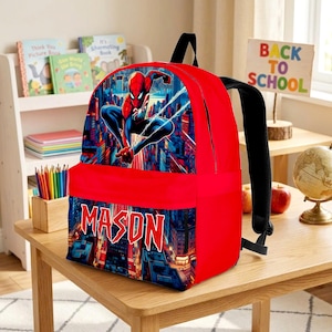 K&ouml;nnte beinhalten: Ein leuchtend roter Rucksack mit einer Grafik des Superhelden Spider-Man in einer dynamischen Pose vor einer Skyline. Die Vordertasche zeigt den Namen MASON in fetten, gezackten wei&szlig;en Buchstaben vor einem detaillierten Stadtbild.