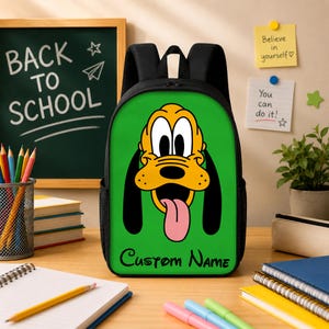 May include: A black backpack featuring a bright green front panel with a large graphic of the cartoon character Pluto, who has yellow fur, long black ears, and a pink tongue. The text Custom Name is printed in black at the bottom of the panel.