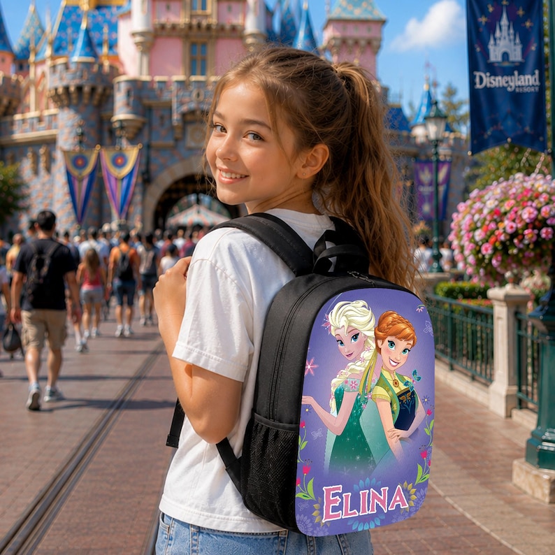 May include: A black backpack featuring a graphic print of the characters Elsa and Anna from Frozen on the back panel. The design includes a purple background with floral accents and the name Elina printed in white letters at the bottom.