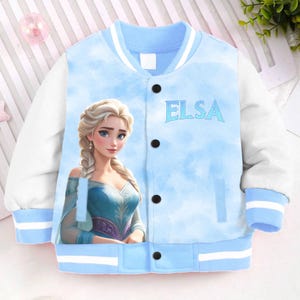 May include: A light blue bomber jacket with white sleeves and a white stripe around the collar. The jacket has a button closure and features a printed image of Elsa from Frozen, along with the text "Elsa".