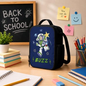 May include: A dark blue rectangular lunch bag featuring a graphic of Buzz Lightyear in a space suit with the text BUZZ in green letters. The bag has a black handle, a side mesh pocket, and a zipper closure, decorated with stars and planet motifs.