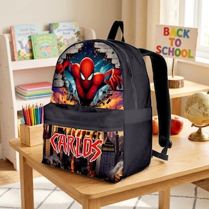 May include: A black backpack featuring a graphic of a red and blue superhero bursting through a brick wall. The lower pocket displays a city skyline at sunset with the name CARLOS printed in a bold, red, jagged font across the center of the bag.