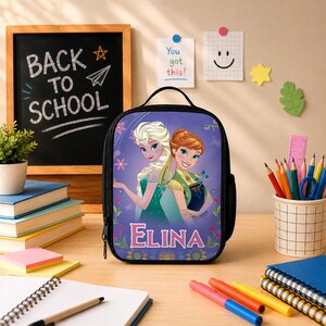 May include: A black lunch bag featuring a graphic print of two animated characters with blonde and auburn hair, surrounded by floral accents and the name ELINA in white lettering. The bag has a top handle and a side mesh pocket for storage.