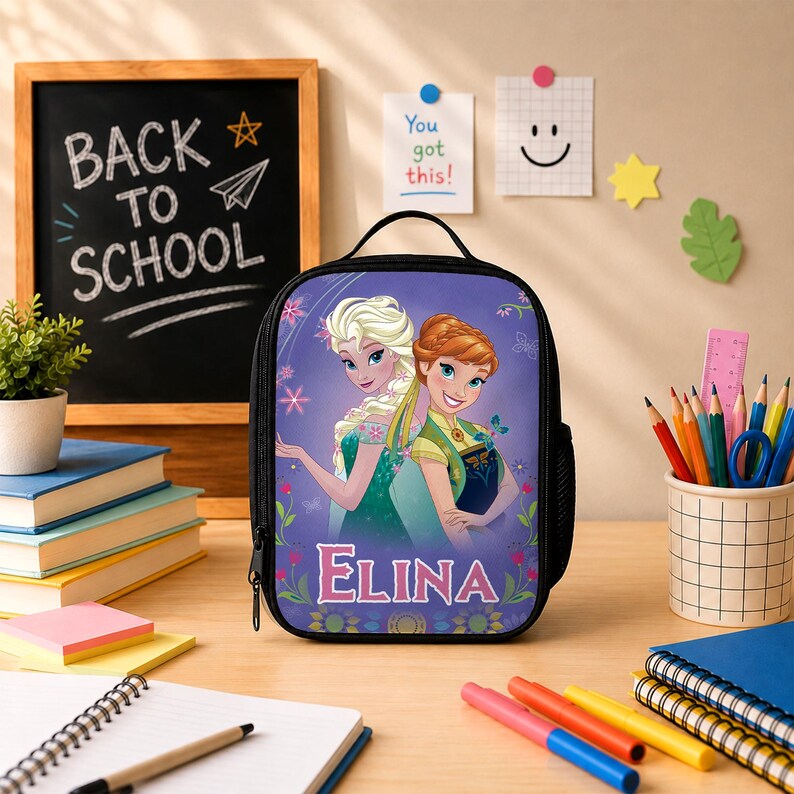 May include: A black lunch bag featuring a graphic print of two animated characters with blonde and auburn hair, surrounded by floral accents and the name ELINA in white lettering. The bag has a top handle and a side mesh pocket for storage.