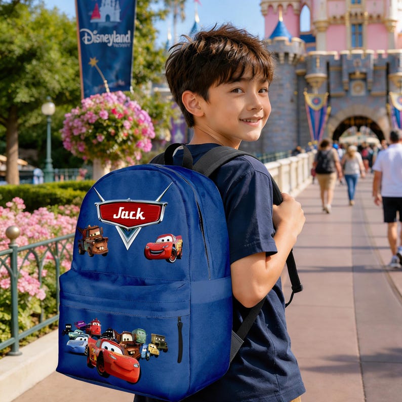 May include: A navy blue backpack decorated with characters from the Cars film, with the name "Jack" displayed on a red sign. The backpack has several images of the Cars characters and a front pocket. The background shows a theme park.
