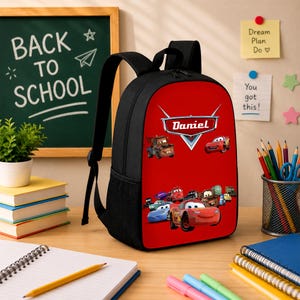May include: A black backpack with a vibrant red front panel featuring a graphic print of characters from the movie Cars and the name Daniel printed in white text. The backpack includes black shoulder straps and side mesh pockets for storage.