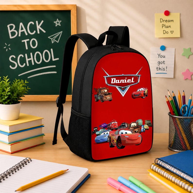 May include: A black backpack with a vibrant red front panel featuring a graphic print of characters from the movie Cars and the name Daniel printed in white text. The backpack includes black shoulder straps and side mesh pockets for storage.