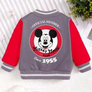 May include: A gray and red baseball jacket with a Mickey Mouse logo on the back. The logo says "Official Member Mickey Mouse Club Since 1955".