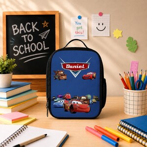 K&ouml;nnte beinhalten: Eine blaue, rechteckige Lunchtasche mit einem Grafikdruck von Charakteren aus dem Film Cars und dem Namen Daniel in einem rot-wei&szlig;en Logostil. Die Tasche verf&uuml;gt &uuml;ber einen schwarzen Griff, einen Rei&szlig;verschluss und eine schwarze Netzseitentasche.