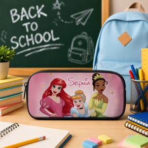 May include: A rectangular pencil case with a pink background featuring illustrations of Ariel, Cinderella, and Tiana. The name Sophia is printed in white script above the characters. The case has a black zip and black trim along the edges.