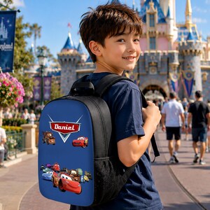 May include: A black backpack featuring a blue front panel with a graphic print of various animated race cars and the name Daniel displayed in a red and white emblem. The bag includes black shoulder straps and side mesh pockets for storage.