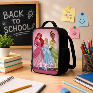 May include: A black lunch bag featuring a graphic print of three princesses in pink, blue, and green gowns with the name Sophia printed in white script. The bag has a top handle, a side mesh pocket, and a zipper closure on the front panel.