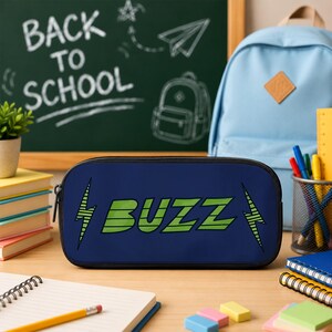 May include: A dark blue rectangular pencil case featuring the word BUZZ in large green striped letters, flanked by two green lightning bolt icons. The case has a black zipper closure and is positioned on a desk with school supplies.