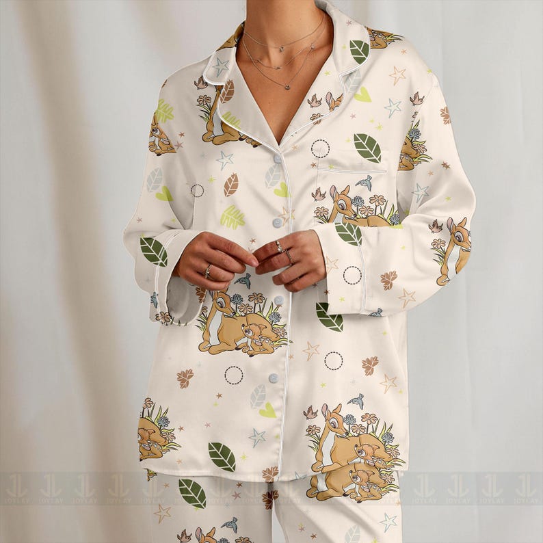 Op de afbeelding: Witte pyjamaset met een Bambi-thema print. De top met lange mouwen en de broek zijn versierd met illustraties van Bambi, bloemen, bladeren en sterren. De top met knopen heeft een reverskraag.