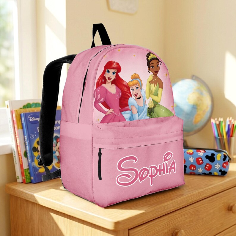 May include: A pink backpack featuring a graphic of three animated princesses on the front pocket area. The name Sophia is printed in a white stylized script font on the lower front pocket. The backpack has black straps and a black top handle.