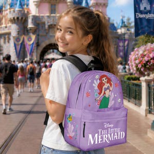May include: A purple backpack featuring Ariel from Disney's The Little Mermaid. The backpack has a front pocket and a graphic of Ariel, seashells, and other sea elements. The words "Disney The Little Mermaid" are printed on the front.
