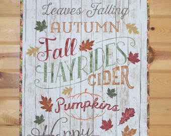 May include: A rectangular wall hanging with a white wood-look background and fall-themed text. The text includes phrases like "Fall is Here," "Harvest," and "Happy Fall Y'all," along with leaf and pumpkin illustrations. The border has a fall leaf pattern.