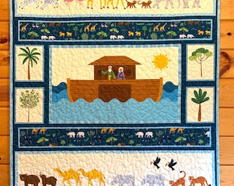 May include: A rectangular quilt featuring a central scene of a wooden ark on blue water with two figures, surrounded by borders of various animals including giraffes, elephants, zebras, camels, and tigers, all set against a cream and blue background.