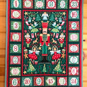 May include: A rectangular fabric advent calendar featuring a central nutcracker illustration surrounded by fairies, Christmas trees, and gifts. The border contains twenty-four numbered squares in red and green with festive botanical patterns.