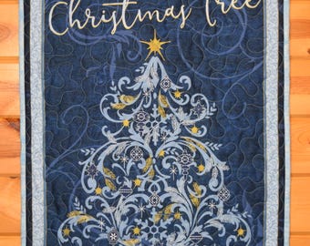 Può includere: Un arazzo da parete trapuntato con sfondo blu navy e un design stilizzato di albero di Natale. L'albero è adornato con elementi decorativi azzurri, dorati e bianchi, e sormontato da una stella dorata. Le parole "Oh, Christmas Tree" sono scritte in corsivo.