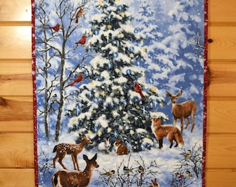 May include: A rectangular fabric wall hanging featuring a winter forest scene with a snow-covered Christmas tree, deer, foxes, rabbits, and red cardinals. The quilt has a red border with white snowflake patterns and hangs from a wooden rod.