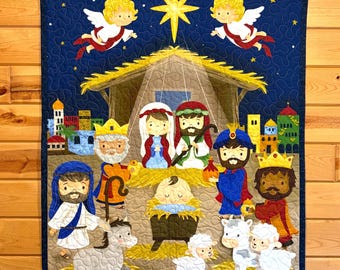 Può includere: Un arazzo da parete trapuntato raffigurante una scena della natività. L'opera presenta uno sfondo blu scuro con una stella e angeli. La scena include figure della sacra famiglia, dei re magi e degli animali, il tutto in stile cartone animato.