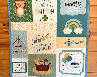 May include: A baby quilt with a patchwork design featuring a lion, a rainbow, and a basket of stuffed animals. The quilt has text that reads "Cute as a Button", "Hello World", and "Welcome Baby". The quilt is trimmed with a blue gingham border.