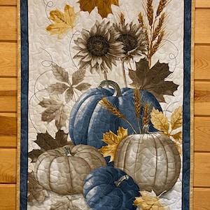 May include: Quilted wall hanging featuring a fall harvest theme. The design includes blue and tan pumpkins, sunflowers, wheat stalks, and autumn leaves in shades of gold, brown, and gray. The border is a dark blue with a gold trim.