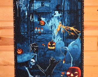 May include: A rectangular fabric wall hanging featuring a dark blue Halloween scene with a haunted house, a witch, black cats, skeletons, and glowing orange jack-o'-lanterns. The quilt has a black border with small orange pumpkin motifs throughout.