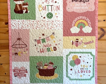 May include: A patchwork baby quilt with various squares featuring foxes, rainbows, and text. The quilt includes phrases like "Cute as a Button," "Hello World," and "Welcome Baby." The color palette is soft, with pinks, greens, and yellows.