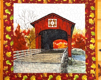 May include: A square fabric wall hanging featuring a central printed image of a red covered bridge surrounded by autumn trees. The center panel has white background quilting and is framed by a border of red fabric with yellow and green leaves.