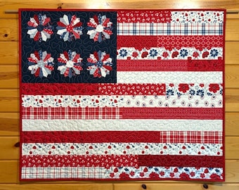 May include: A handcrafted quilt depicting the American flag. The quilt features a navy blue field with six starburst designs. The stripes alternate between red and white patterns, including floral and plaid designs. The quilt is displayed on a wooden wall.