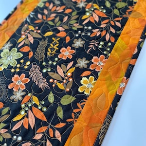 May include: Quilted table runner with a black floral design in orange, yellow, and green. The runner has an orange border with a leaf pattern. The runner is approximately 30 cm wide.
