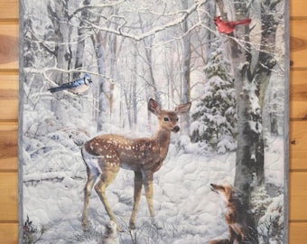 May include: A rectangular fabric wall hanging featuring a winter forest scene with a spotted deer, a fox, a squirrel, a blue jay, a cardinal, and an owl. The quilt has a gray border and displays a snowy landscape with bare trees and pine branches.