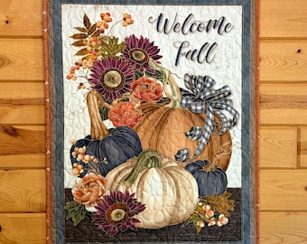 May include: A rectangular fabric wall hanging featuring a quilted design with the text "Welcome fall". The image displays a collection of pumpkins in orange, white, and dark blue, accented with sunflowers, orange flowers, and a blue gingham bow.