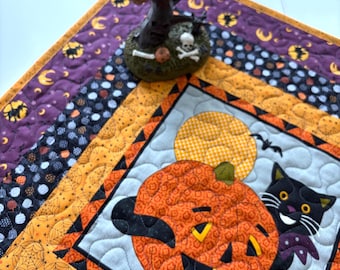 May include: A square quilted fabric mat featuring a central panel with a black cat and a jack-o'-lantern against a light gray background, surrounded by orange and purple borders with spiderweb and moon patterns, topped with a witch figurine.