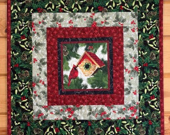 Può includere: Tovaglietta natalizia trapuntata quadrata con un'immagine centrale di un cardinale rosso e una casetta per uccelli. Il design presenta strati di tessuti rossi, verdi e bianchi con motivi di agrifoglio, pigne e fiocchi di neve. Un bordo rosso incornicia il design.