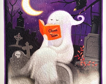 May include: A rectangular fabric wall hanging quilt featuring a white ghost sitting on a tombstone in a graveyard at night, reading an orange book titled Ghost Tales. The scene includes a yellow crescent moon, a black crow, and purple sky.