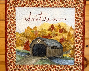 May include: A square fabric wall hanging featuring a quilted scene of a covered bridge surrounded by autumn trees. The text "adventure AWAITS" is printed in dark red script and block letters. The border has a pattern of small orange leaves.